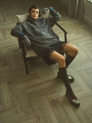Model seated in an armchair wearing an oversized dark grey hoodie sweater, fringe trim skirt, knee socks, and black loafers.