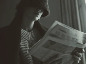 Model wearing a low-brimmed hat obscuring eyes and reading a newspaper in moody lighting for Brunello Cucinelli.