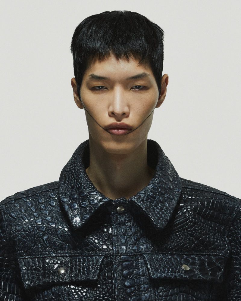 Close-up portrait of a young Asian male model with dark hair wearing a dark blue crocodile-print jacket against a white background.