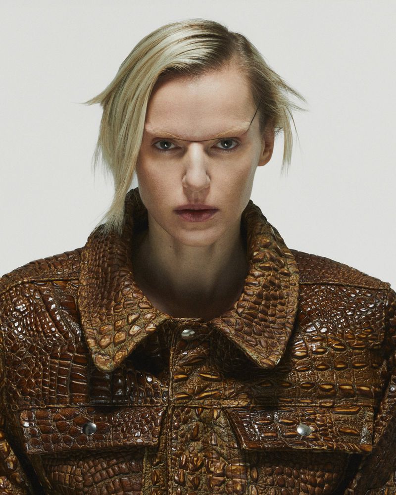 Close-up of a fair-skinned adult woman with short blonde hair wearing a brown reptile-patterned jacket against a white backdrop.