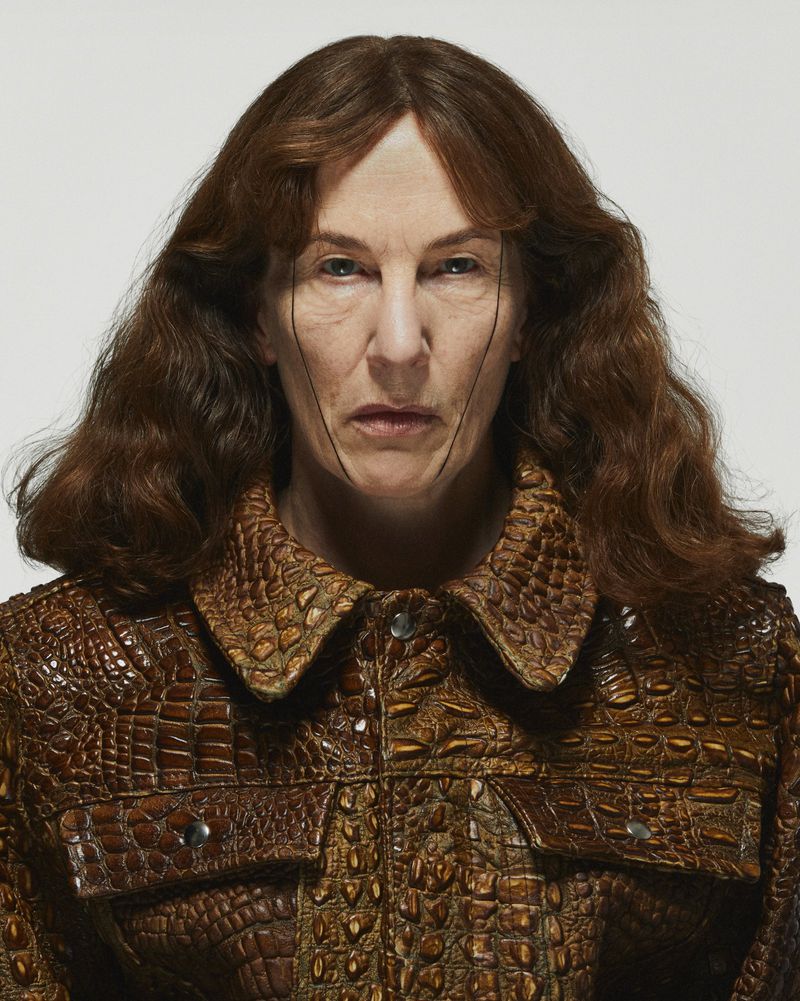 Portrait of an adult woman with brown hair wearing an alligator-skin-textured jacket, standing indoors against a white background.