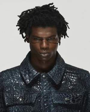Portrait of a black man with dreadlocks wearing a textured dark blue embossed jacket against a plain light grey background.