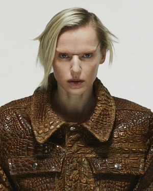 Model Elana Mityukova wearing a brown croc-embossed leather jacket and wire bridge glasses against a light grey seamless backdrop.