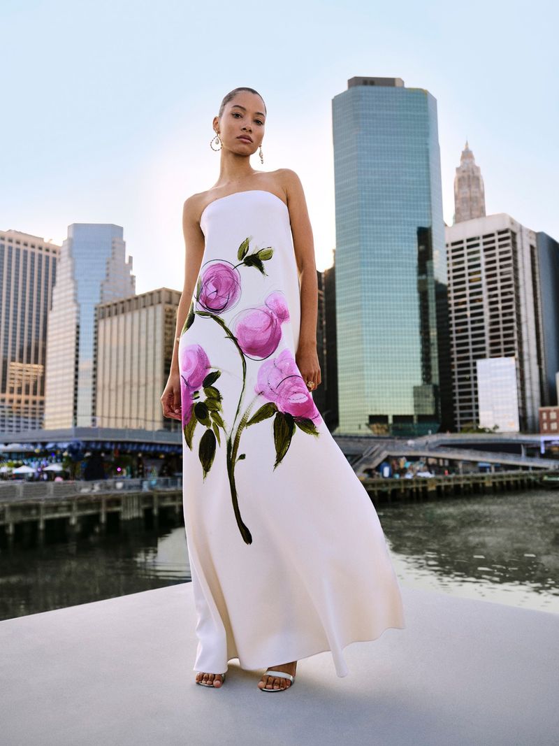 Woman in a strapless gown with pink floral design poses against a backdrop of city buildings and a blue sky at daytime.