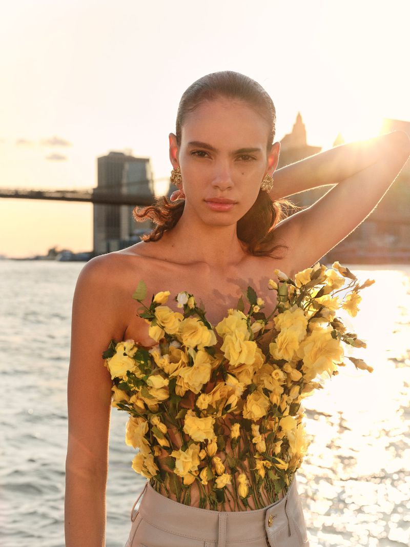 Young woman poses in a strapless floral top against a backdrop of the Brooklyn Bridge and a sunlit body of water in an outdoor setting.