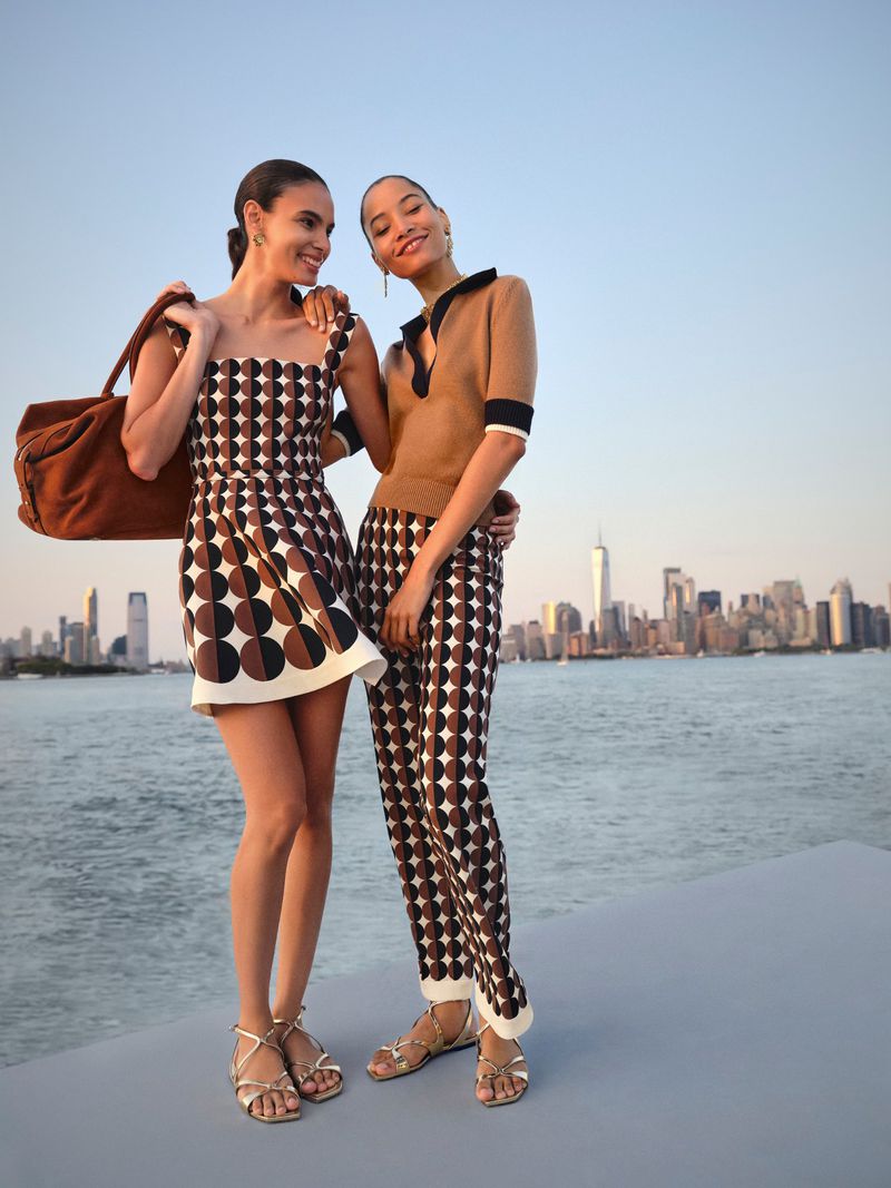 Two women pose in patterned dresses with a cityscape backdrop. One carries a brown duffel bag on her shoulder, standing on a rooftop.