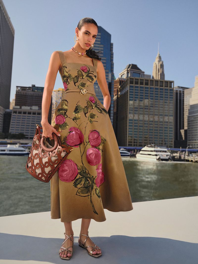 Adult female fashion model wearing floral dress and holding designer handbag poses against a modern cityscape backdrop on sunny day.