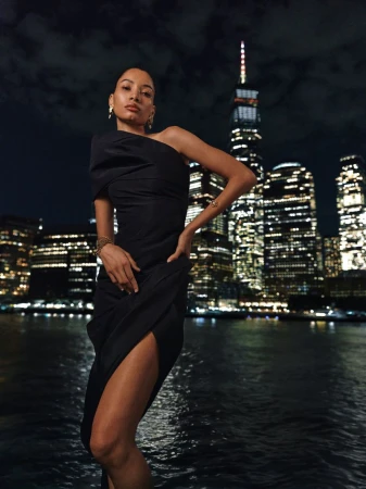 Model wearing a black one-shoulder dress posing against a night cityscape with illuminated skyscrapers across water