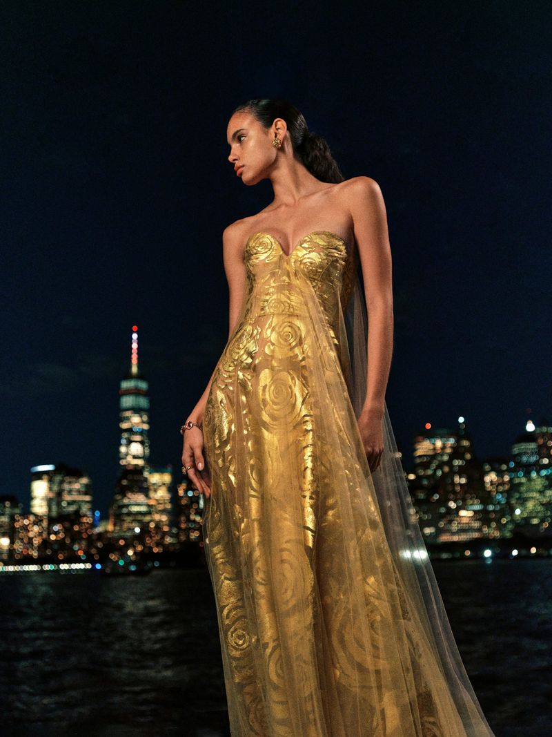 Adult female model in golden gown posing with city skyline at night. Elegant strapless dress with sheer shawl and swirling pattern.