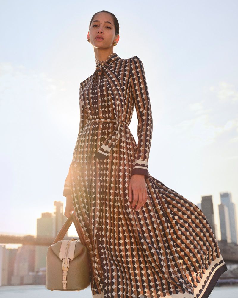 Woman in geometric print dress with a handbag stands against a city skyline backdrop on a sunny day; fashion outdoor editorial shot.