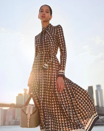 Woman in geometric print dress with a handbag stands against a city skyline backdrop on a sunny day; fashion outdoor editorial shot.
