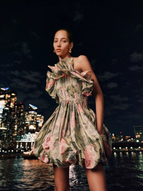 Woman in floral mini dress posing outdoors at night with city lights reflecting in the water in the background, fashion editorial shot.