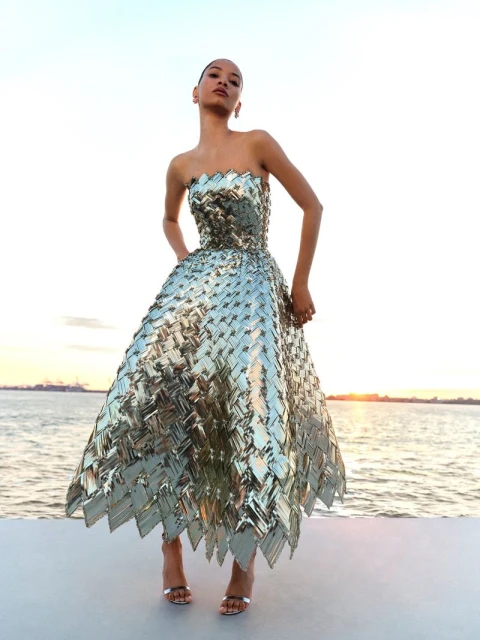A woman poses in a shimmering, strapless gown with a full, textured skirt against a soft sky and ocean backdrop at sunset, wearing heels.