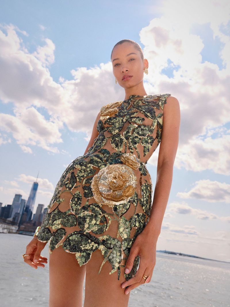 An adult female model poses in a floral embellished tan mini dress with a bodycon silhouette in front of a city waterfront under a blue sky.