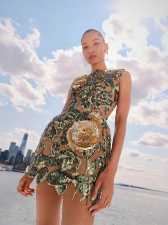 An adult female model poses in a floral embellished tan mini dress with a bodycon silhouette in front of a city waterfront under a blue sky.