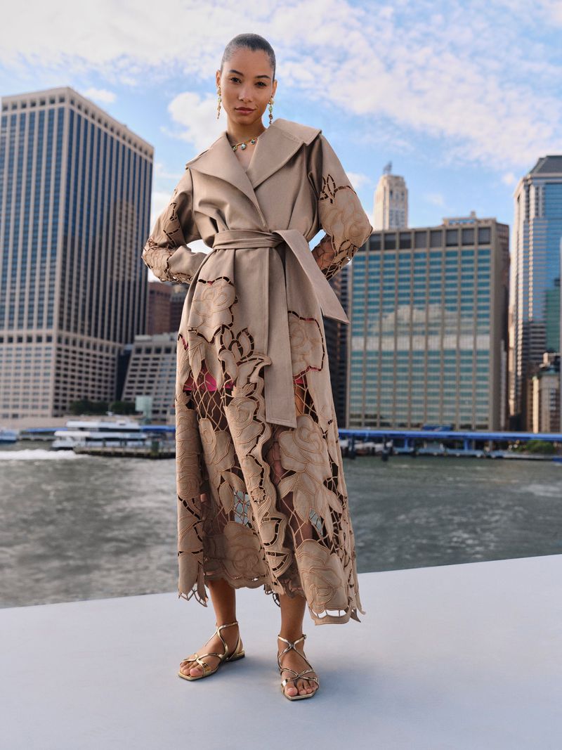 Model wearing a tan belted coat with laser-cut floral patterns standing outdoors with city skyline background.