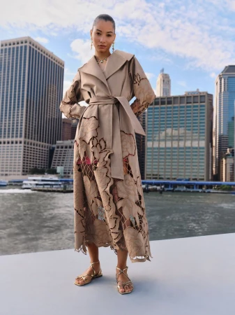 Model wearing a tan belted coat with laser-cut floral patterns standing outdoors with city skyline background.