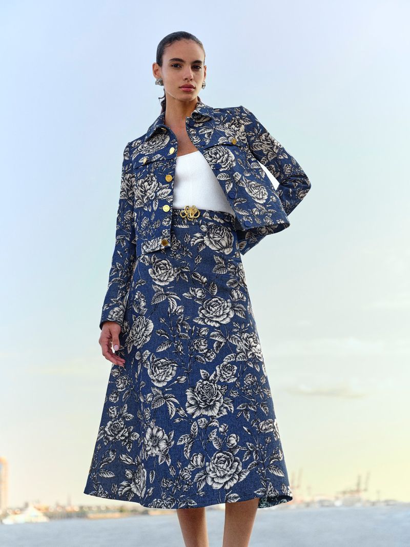 A woman poses outdoors wearing a floral print denim jacket and a-line skirt, accessorized with a white top and gold belt and earrings.