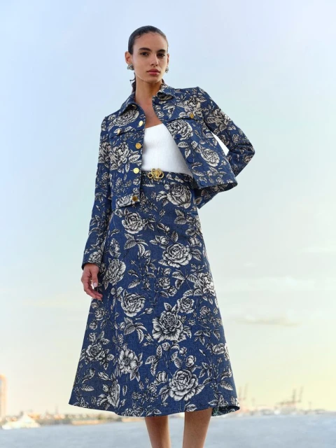 A woman poses outdoors wearing a floral print denim jacket and a-line skirt, accessorized with a white top and gold belt and earrings.