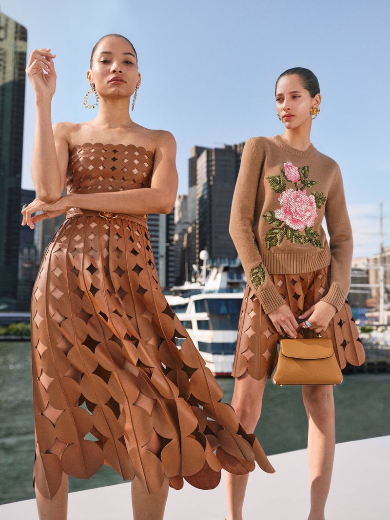 Two women pose in matching outfits on a sunny day, featuring a brown laser-cut skirt and top set against a city and water backdrop.