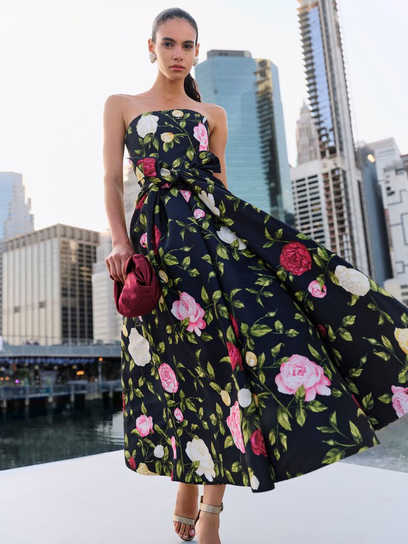 Woman in a floral strapless dress holding a burgundy purse, posing on a rooftop with a cityscape in the background during daylight.