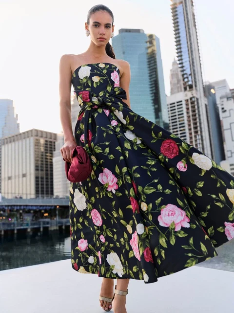 Woman in a floral strapless dress holding a burgundy purse, posing on a rooftop with a cityscape in the background during daylight.