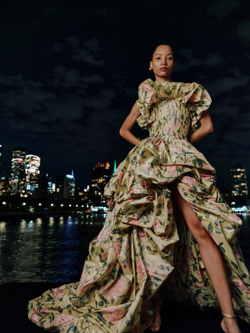 Woman in floral gown poses at night, city skyline in background. The dress has ruffled details and a dramatic skirt with a high slit.