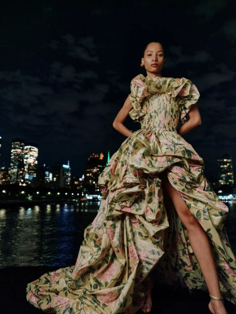Woman in floral gown poses at night, city skyline in background. The dress has ruffled details and a dramatic skirt with a high slit.