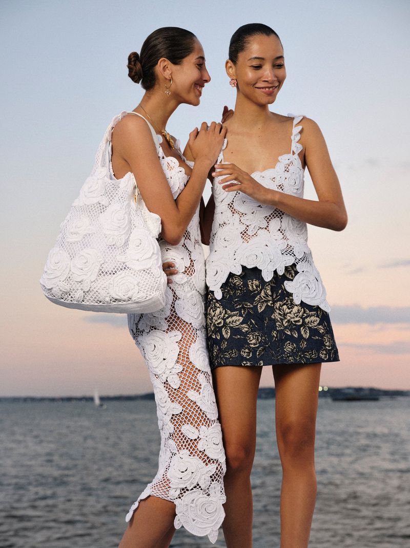 Two women pose outdoors wearing matching white floral-applique tops; one wears a floral miniskirt and the other a matching full-length skirt.
