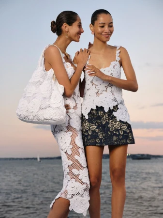 Two women pose outdoors wearing matching white floral-applique tops; one wears a floral miniskirt and the other a matching full-length skirt.
