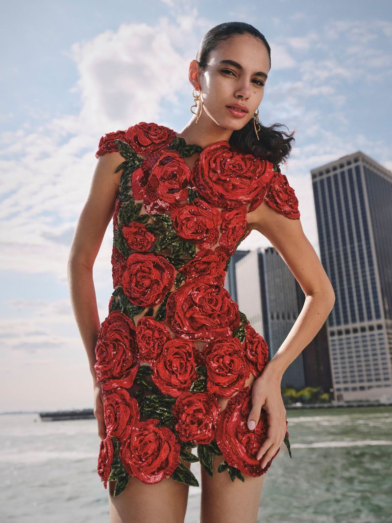 A woman in a red rose-covered mini dress poses outdoors with a city and sky background, showcasing a striking fashion-forward floral design.