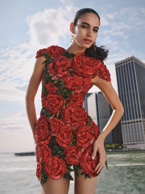 A woman in a red rose-covered mini dress poses outdoors with a city and sky background, showcasing a striking fashion-forward floral design.