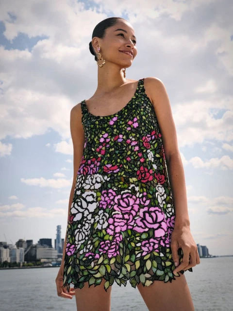 A woman poses in a floral dress with a city skyline backdrop under a partly cloudy sky, smiling and looking off-camera.