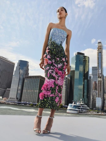 Fashion model in floral-print dress poses outdoors with skyscrapers and a river as backdrop, wearing metallic high-heel sandals and jewelry.