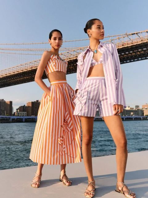 Two women in matching striped summer outfits pose near the Brooklyn Bridge with the city skyline and water in the background.