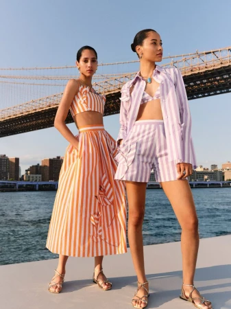 Two women in matching striped summer outfits pose near the Brooklyn Bridge with the city skyline and water in the background.