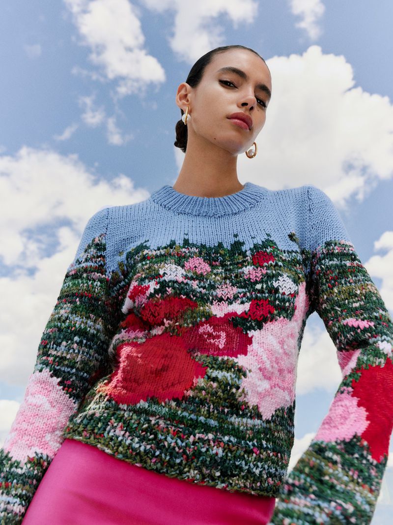Model wearing a blue knit sweater with floral landscape pattern against a bright blue sky with clouds.