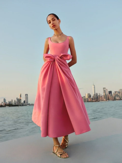 A stylish adult female stands outdoors in a pink formal gown with a large bow, against an urban skyline and blue sky backdrop.
