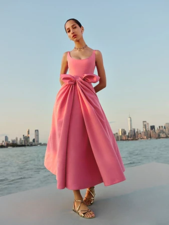 A stylish adult female stands outdoors in a pink formal gown with a large bow, against an urban skyline and blue sky backdrop.