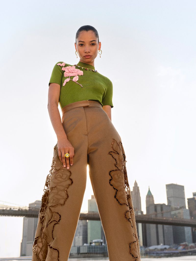Fashion photo of an adult woman posing outdoors in an olive-green top and tan wide-leg trousers with floral appliques, city skyline.