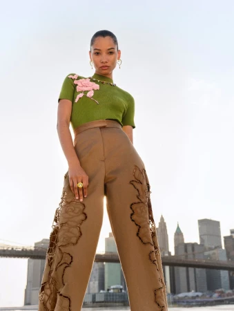 Fashion photo of an adult woman posing outdoors in an olive-green top and tan wide-leg trousers with floral appliques, city skyline.
