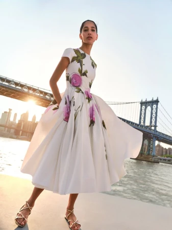Woman in a floral dress stands with hands on hips against the backdrop of a bridge and city buildings under a clear, bright sky.