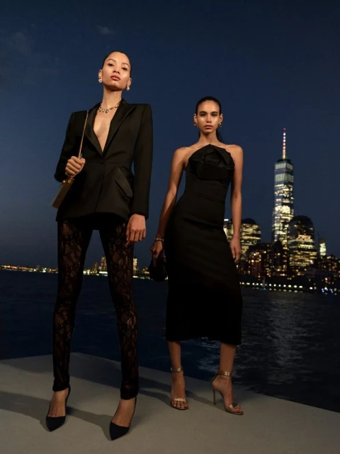 Two women pose in evening wear with a city skyline at night. One wears a blazer and lace pants, the other a strapless dress.