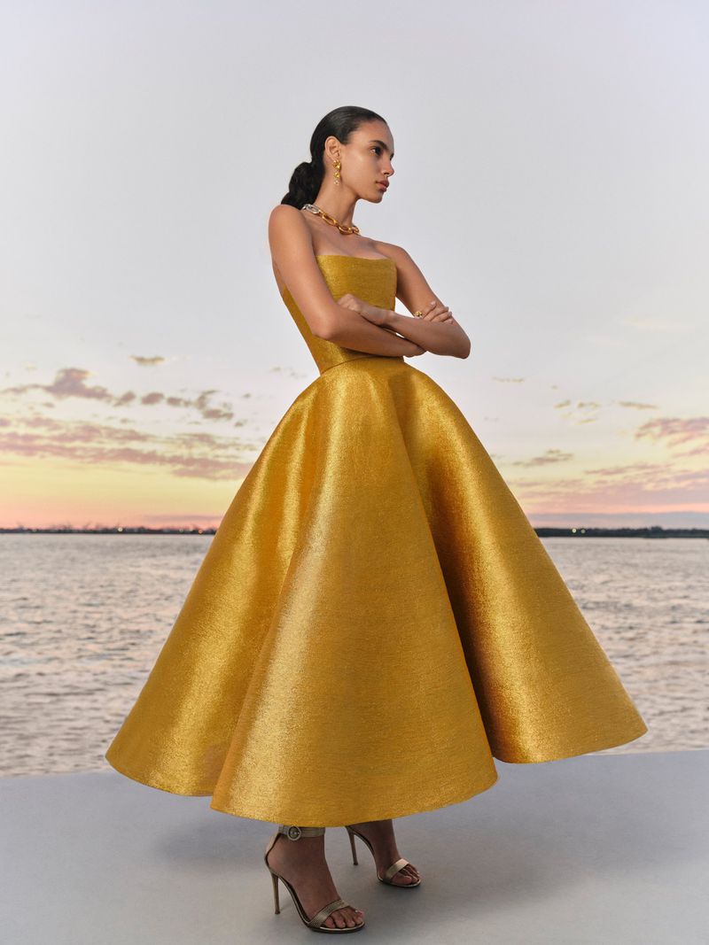 Model in a strapless golden gown with a voluminous flared skirt posing against an outdoor water and sunset sky backdrop.