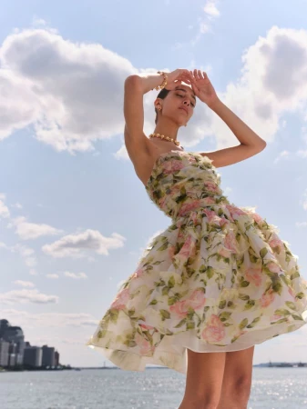 Model in strapless floral dress with arms raised against a bright cloudy sky and water backdrop