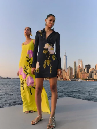 Two fashion models pose in dresses with floral prints on a city harbor with skyscrapers in the background on a sunny day.