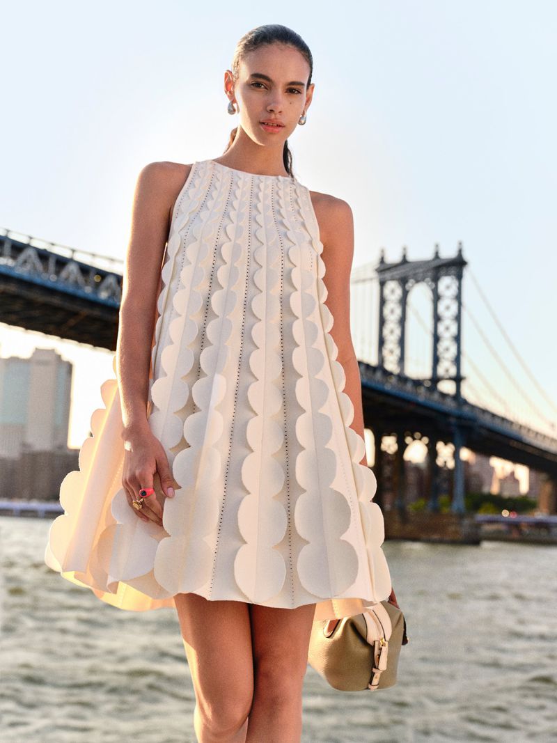 Fashion shoot of a woman wearing a cream-colored, above-the-knee dress with circular designs, posing in front of a bridge and river.