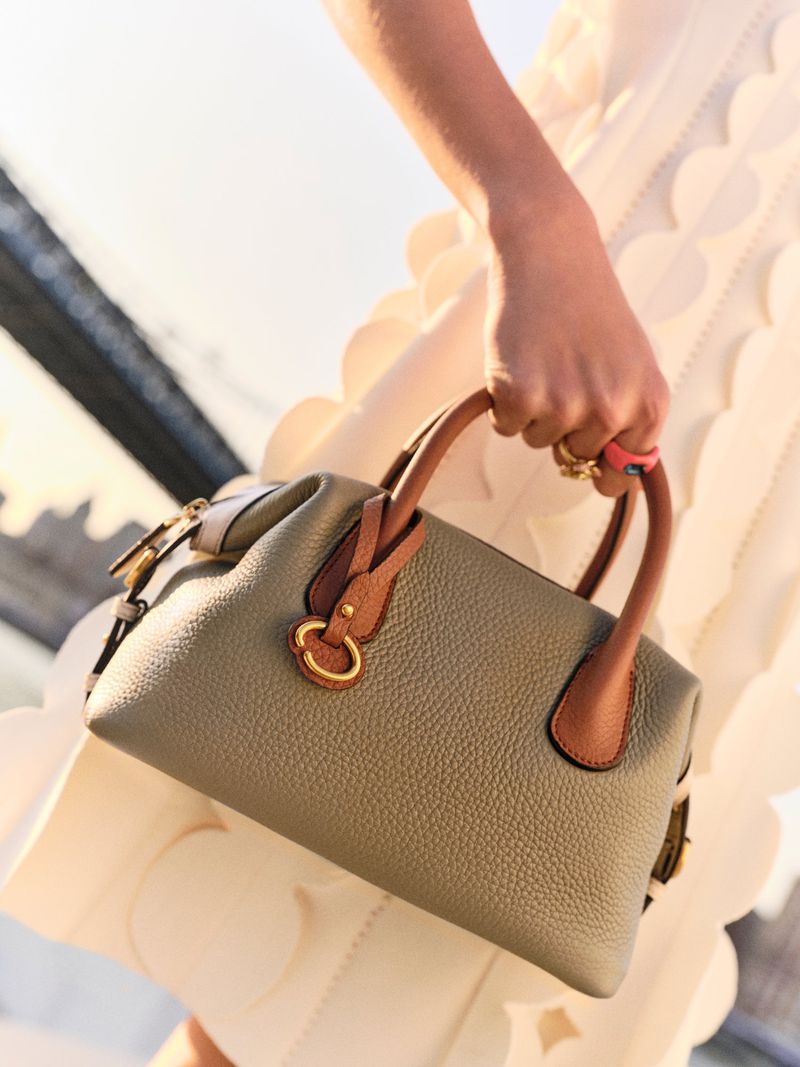 Close-up of a woman carrying a gray leather satchel handbag with tan handles, wearing a white textured dress, outdoors in soft lighting.