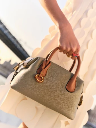 Close-up of a woman carrying a gray leather satchel handbag with tan handles, wearing a white textured dress, outdoors in soft lighting.