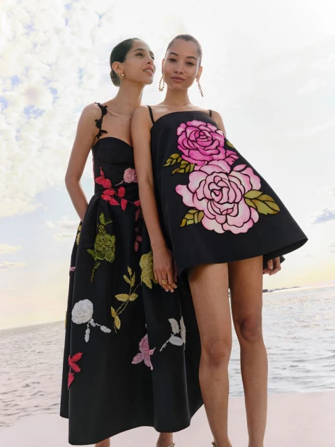 Two women pose in elegant black dresses with floral embroidery against a bright sky and water backdrop for a fashion editorial.